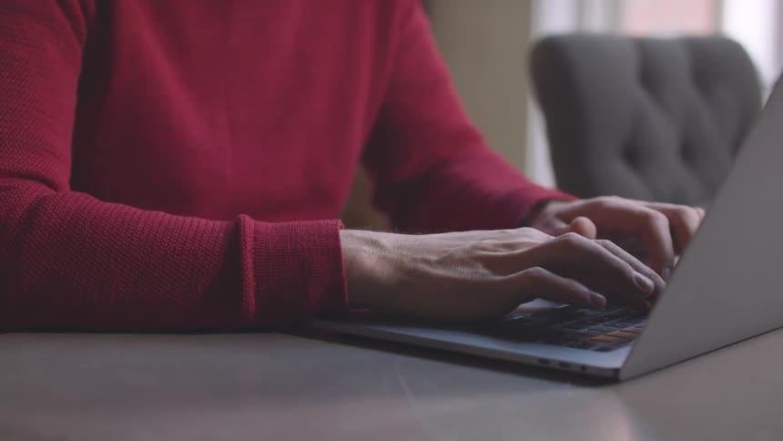 A woman wearing a red sweater types on a laptop keyboard at a desk in a dimly lit office