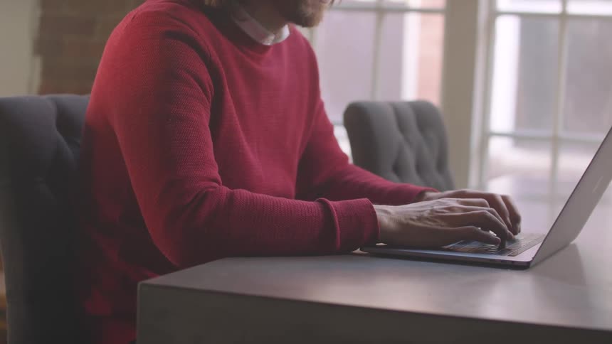 Man sitting at a table near windows typing on a laptop computer