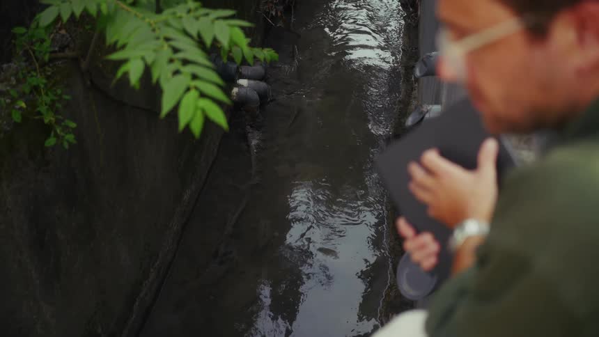 A person stands beside a muddy drainage canal observing water flow from multiple pipes under overhanging leaves and concrete walls