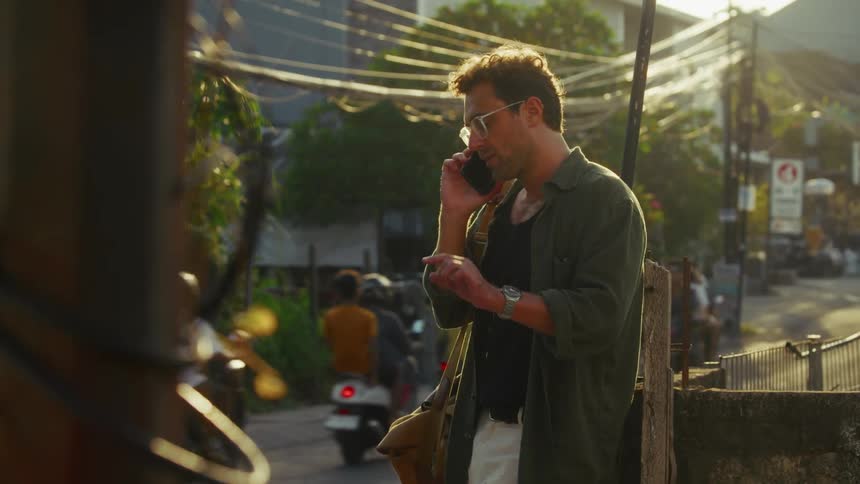 A person stands on a street in a dark green shirt talking on a mobile phone with power lines and a mix of buildings and foliage in the background