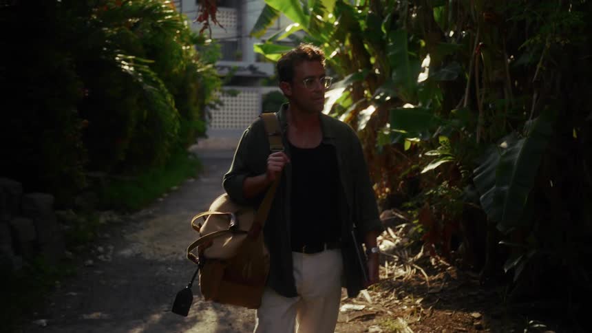 Man carrying a large bag checks his wristwatch while walking away from a building along a path between trees on a sunny day