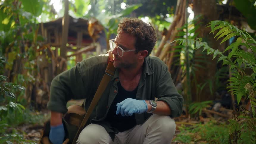 A person wearing blue gloves handles a sample in a plastic container while crouching in a dense tropical setting near a rural settlement