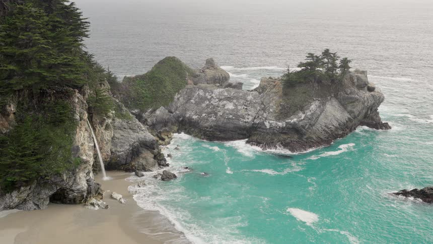 An aerial view of a waterfall cascading onto a sandy beach cove with turquoise waters surrounded by rocky cliffs and lush vegetation under an overcast sky