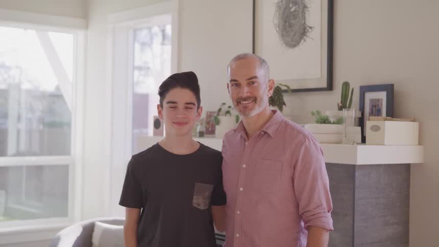 A father and son are standing side by side in a living room smiling at the camera