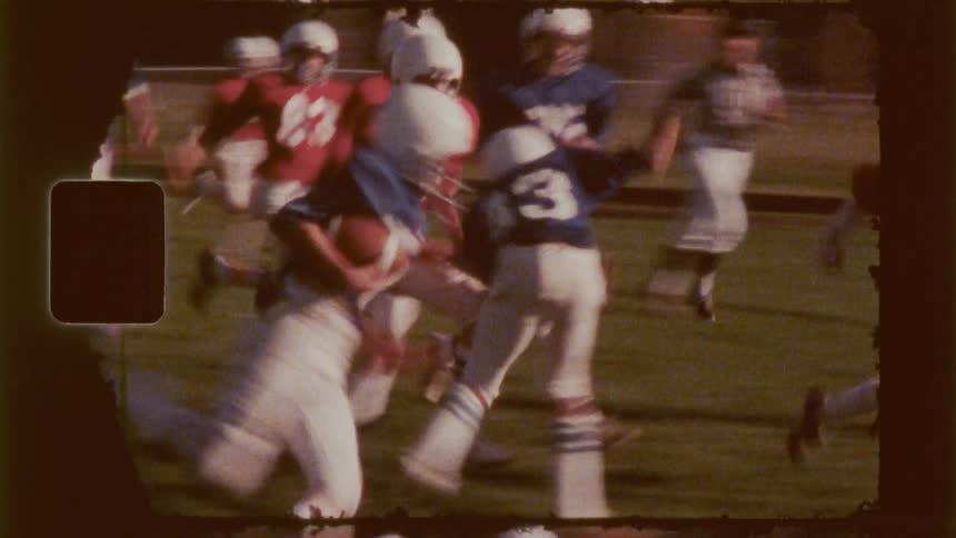 A vintage slightly blurry film shows a football play in action on a grassy field with players in blue and red uniforms
