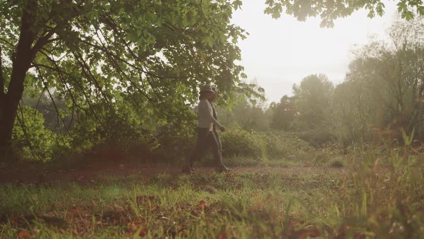 Dressed for fall a man and woman hold hands while walking across a sundappled grassy field surrounded by trees enjoying a moment outdoors together