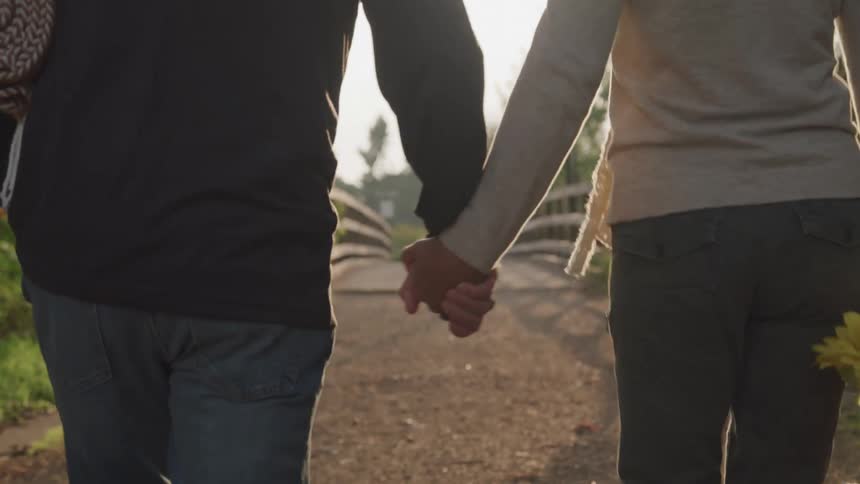 Two people walk hand in hand down a sunlit path one carrying a rolled up mat and the other a bouquet of sunflowers