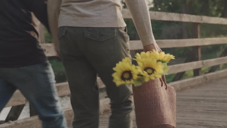 Woman carrying flowers in a bag walks hand in hand with a man as they cross a wooden bridge during the day