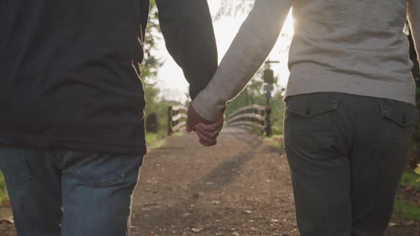 A couple walks hand in hand along a path towards a small bridge in the distance