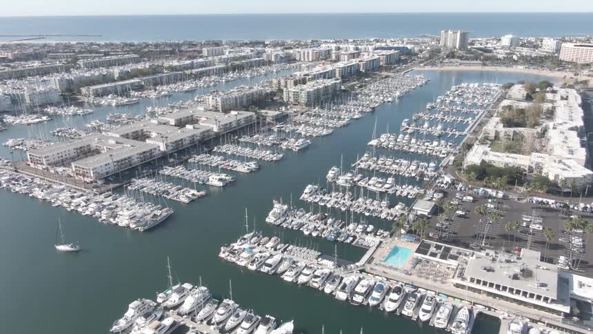 An aerial view shows a densely packed marina with numerous white boats docked in neat rows surrounded by buildings and a parking lot