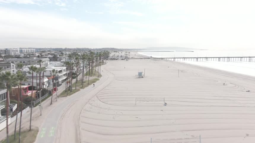 A long sandy beach with a pier extending into the ocean lined with palm trees and a pathway where people are walking and biking with a city skyline visible in the distance under a bright overcast sky