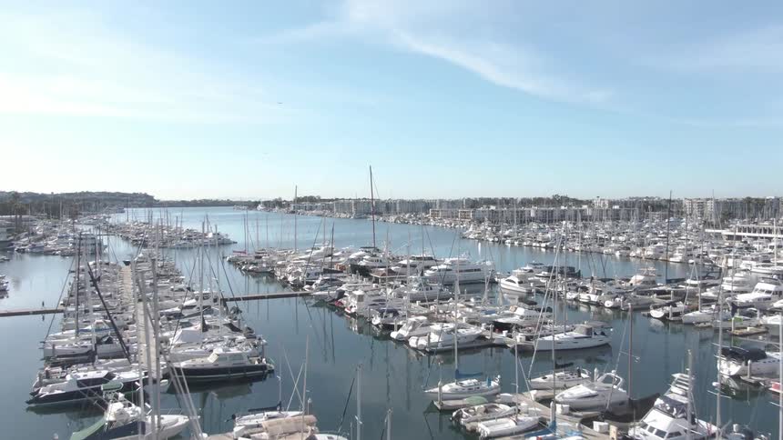 This aerial shot looks down upon a large crowded marina where countless sailboats and yachts are moored at docks on calm water during the daytime