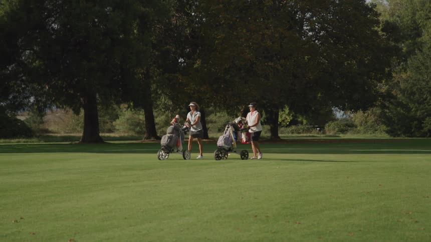 Two female golfers push carts carrying their golf clubs across the green on a summer day