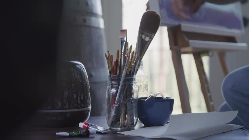 A persons hand reaches into a glass jar filled with paintbrushes on a table alongside paint tubes a blue mug and paper with an easel visible in the background