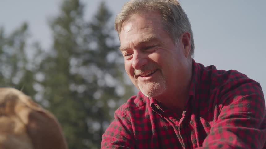 Older man smiles while petting his dog outdoors on a sunny day