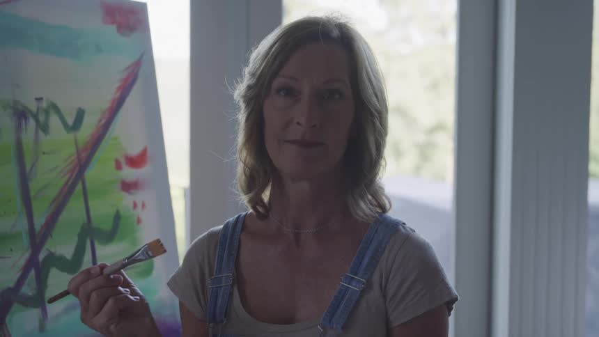 A painter with shoulder length blonde hair stands holding a paintbrush in front of a partially painted canvas