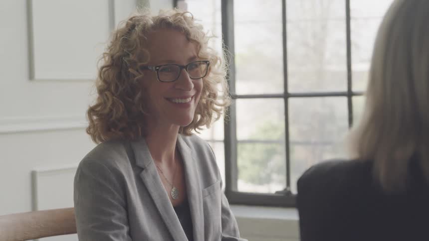 A businesswoman with curly blonde hair and glasses is engaged in conversation with her coworker