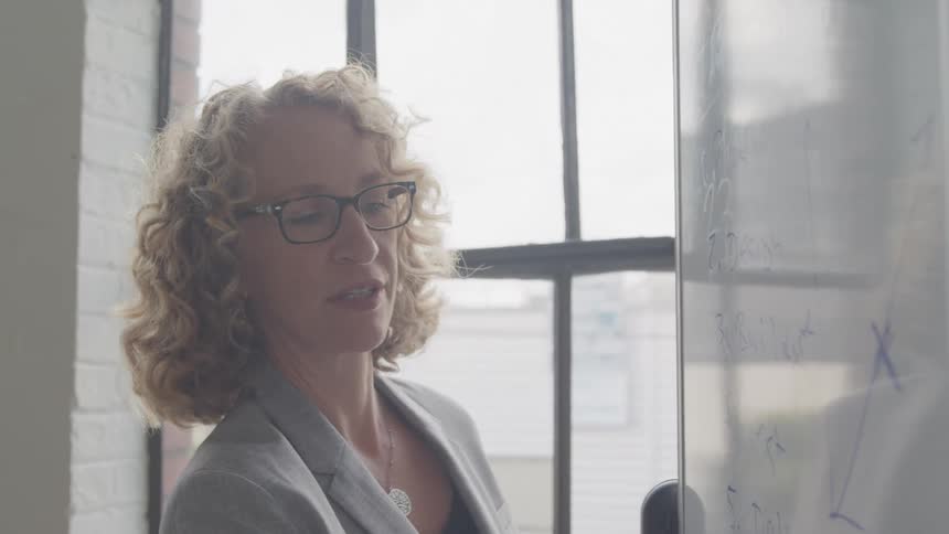 A woman with curly hair wearing glasses and a blazer writes on a whiteboard near a window in a brightly lit room