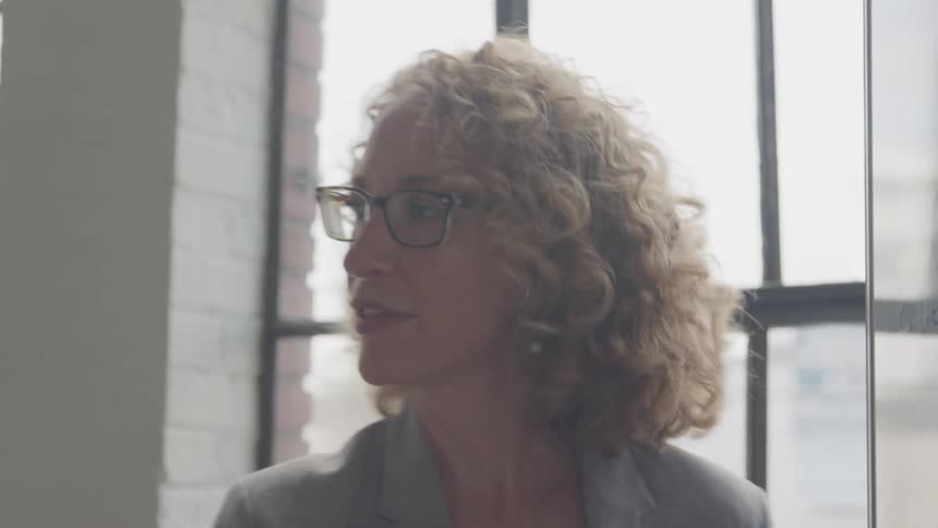 The blonda haired woman with the glasses is writing something on a white board and talking to someone behind her