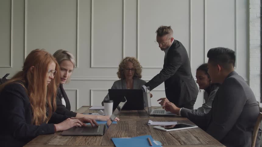 A group of six people are gathered around a wooden table in a meeting with some looking at laptops and others engaged in discussion