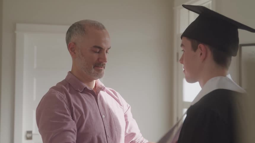 A father proudly adjusts his sons graduation cap and toga in a brightly lit room at home near a window