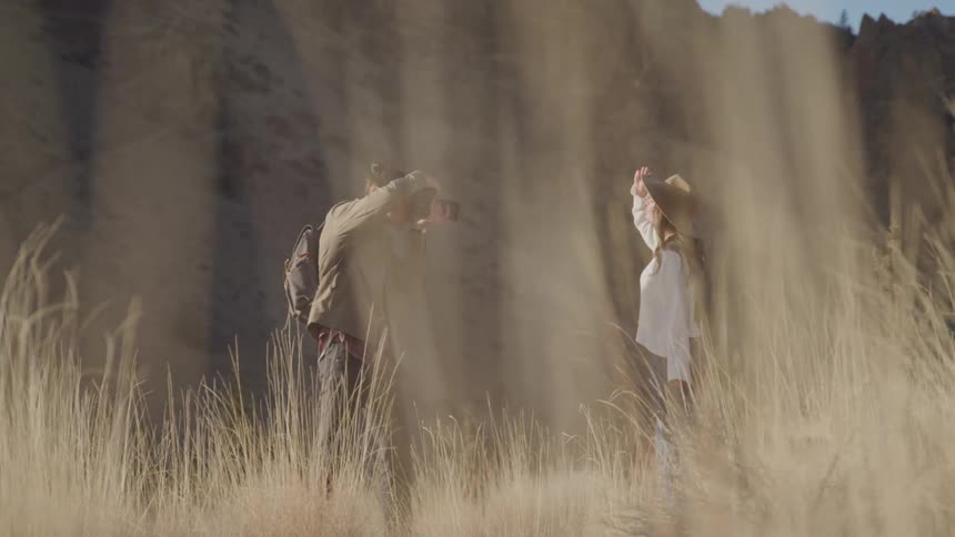 A photographer partially obscured by tall grass takes pictures of a woman posing in front of a rocky backdrop