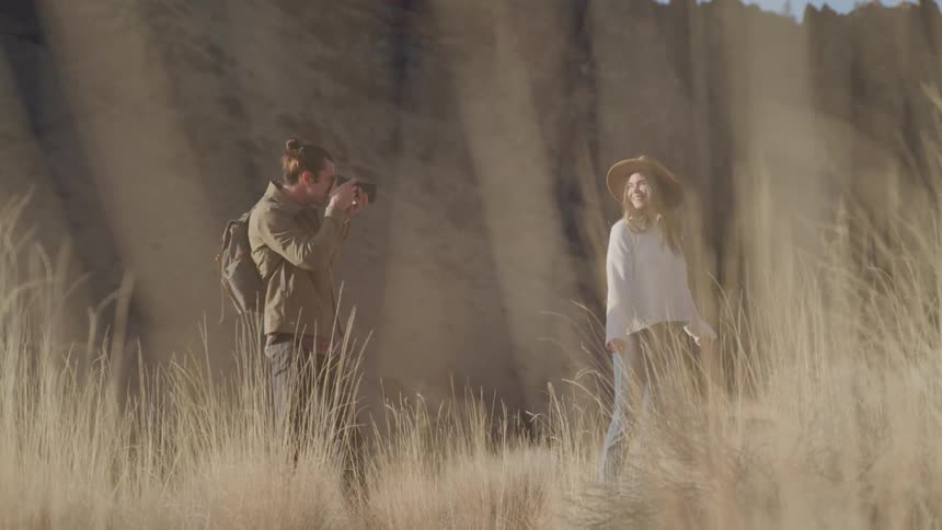 A woman in a white sweater and hat poses for a photographer amidst tall dry grass
