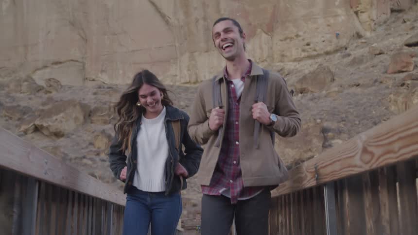 A man and a woman walking across a wooden bridge they are smiling while the man is wearing a backpack
