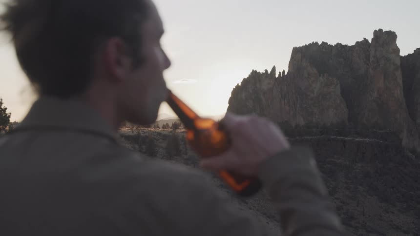 A man is standing outside drinking a beer and looking at a mountain