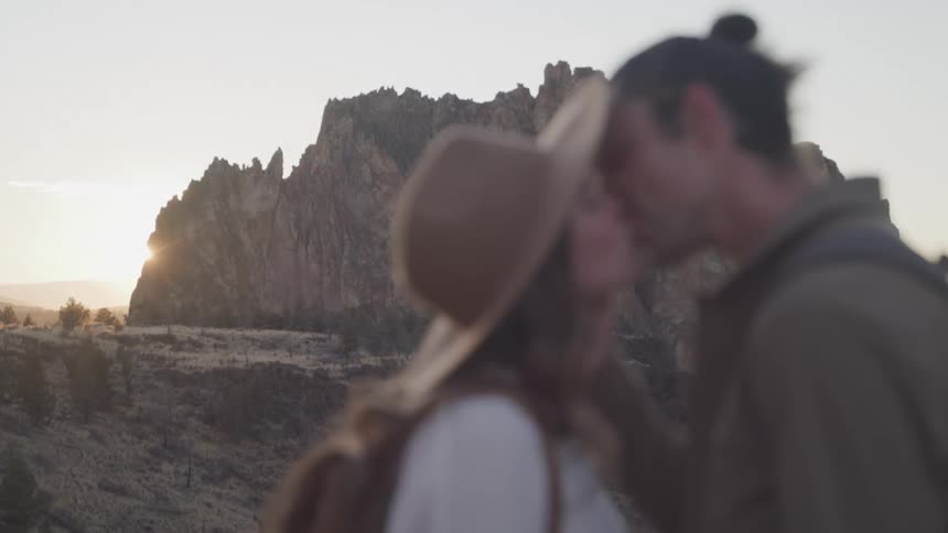A man and a woman kiss each other in front of a jagged mountain with the sun setting behind a desert landscape