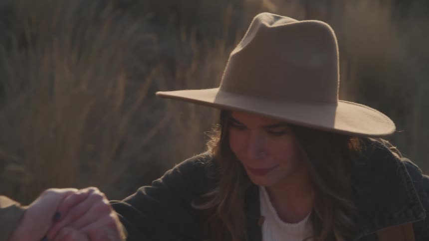 A man extends his hand to help a woman up a hill she wears a sun hat and has long hair with warm sunset light in the background