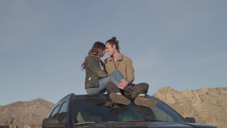 A young couple affectionately sit atop a car in the middle of a desert area