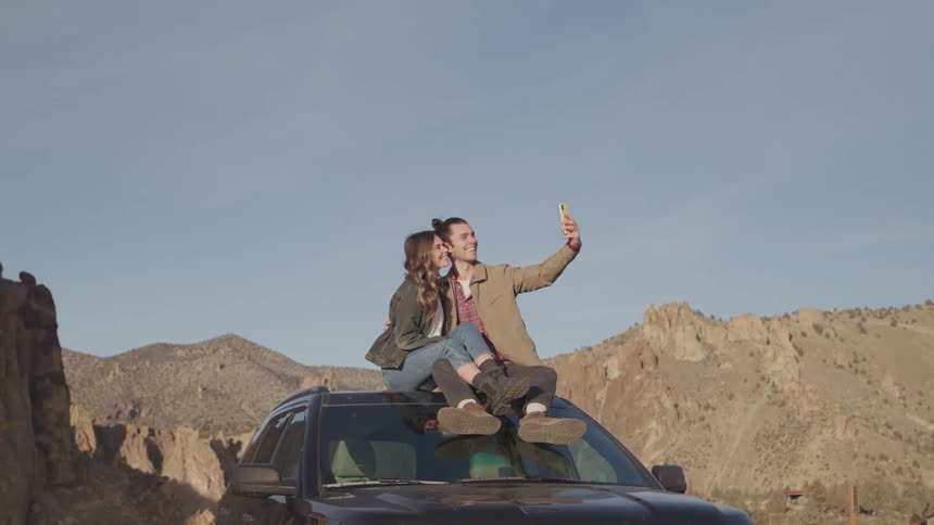 A young couple sits on the roof of a car taking a selfie with a phone surrounded by a desert landscape in daytime