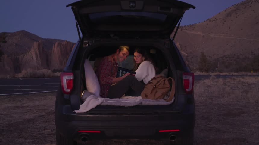 A couple sits in the back of an suv at dusk using a tablet with a scenic mountainous landscape in the background