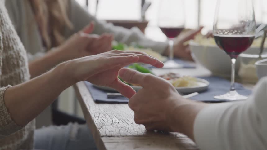 A closeup of people holding hands in prayer around a table set for lunch with wine food and plates in a bright room