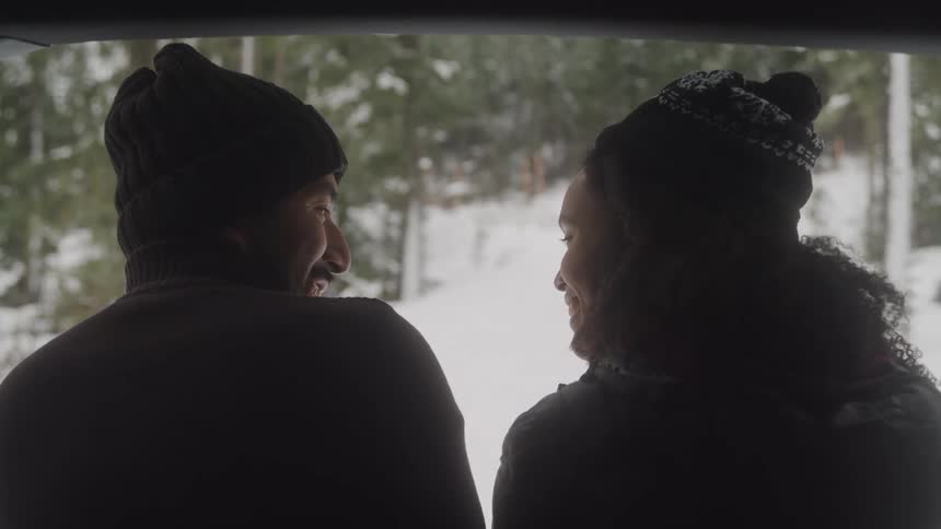 Two people in winter hats sit next to each other facing each other with a snowcovered forest visible outside