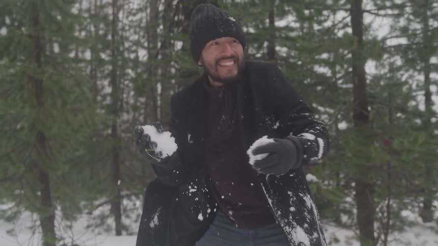 A man in a black coat and beanie smiles as he prepares to throw snowballs in a snowy forest