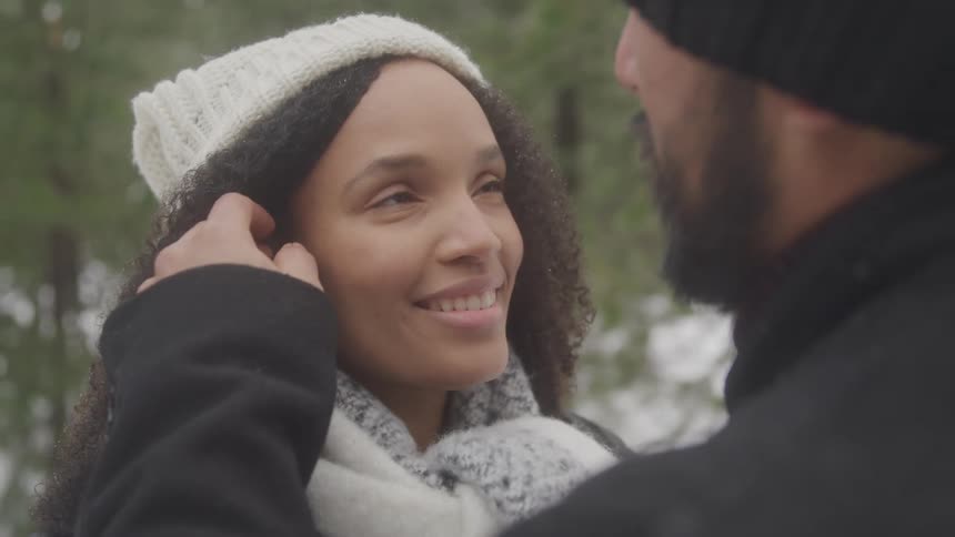 A woman with dark curly hair and a white knit hat smiles lovingly at a man in a snowy outdoor setting where he is gently touching her hair