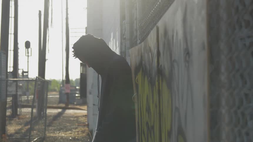 Black man loiters against a graffitied fence