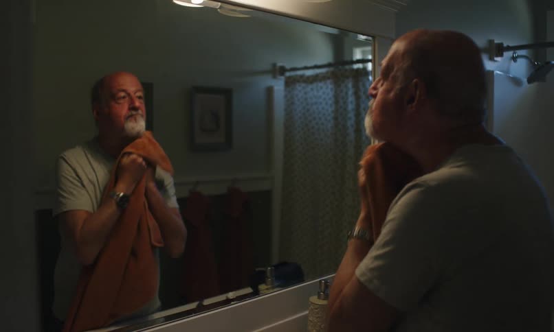 A man with a gray beard dries his face with a towel in a bathroom mirror