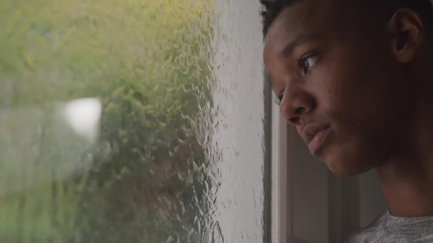 Sad teenage boy leaning on window looking at rain running down the glass