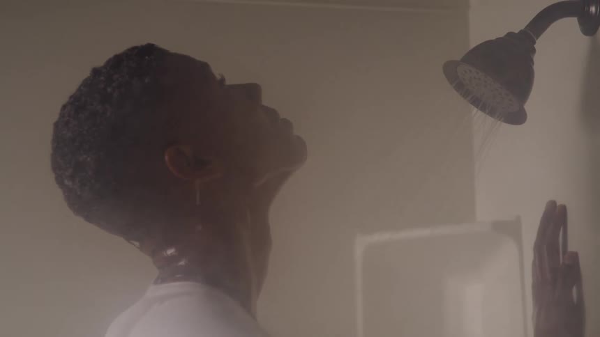 The black man is standing in the shower with his white shirt on just letting the water run over him