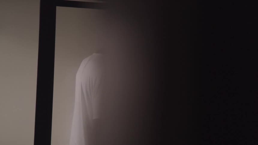 A black man in a white tshirt is in a clean bathroom urinating with his left hand on the wall and his other holding himself while a camera outside the window films him candidly