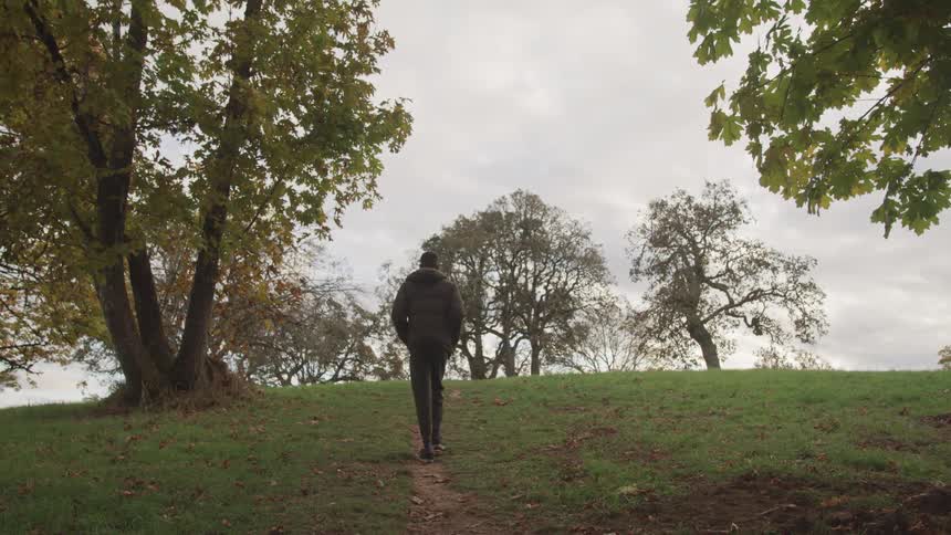 A man walks along a path through wooded area with tall trees framing the scene under a cloudy sky