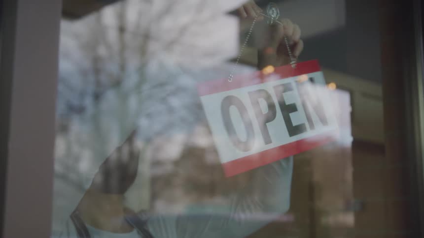 Female employee wearing a face mask turning closed sign around to open from inside window