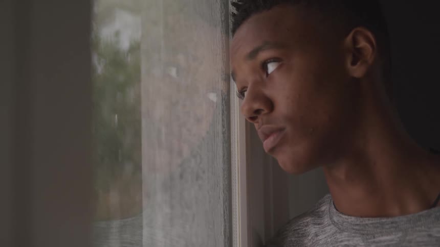 A teenage boy is looking out the window when its raining