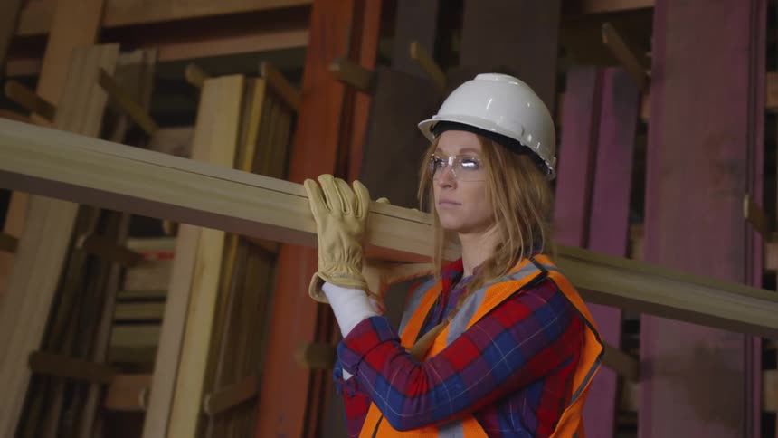 Woman uses both hands to balance lumber on her shoulder as she carries it
