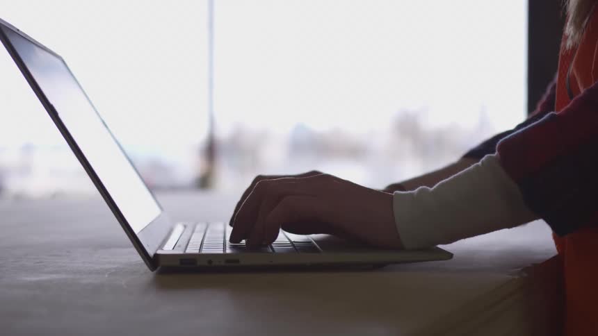 A womans hands typing on a laptop keyboard with a bright blurred window in the background