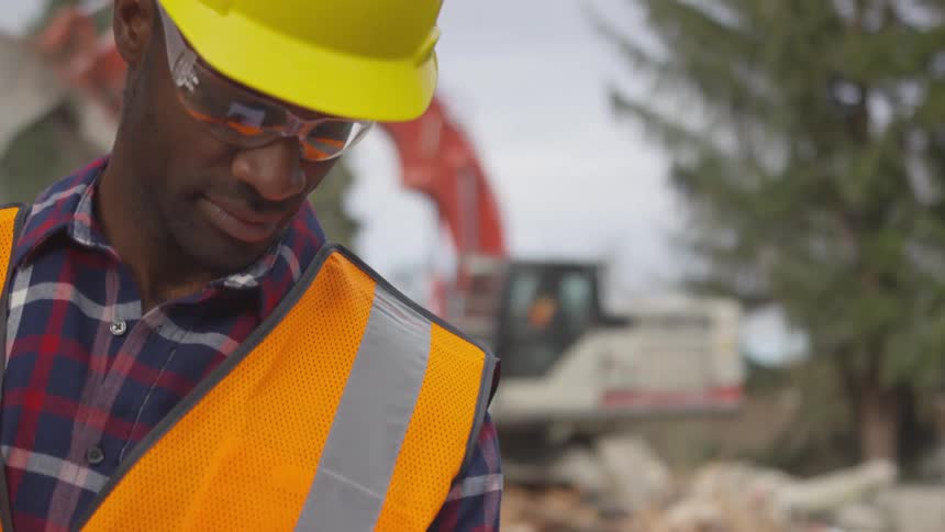 The black man who is wearing the safety glasses and vest along with the hard hat is looking down at something while the back hoe is working behind him