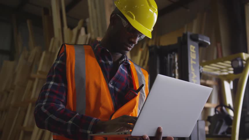 Man in a hardhat types on a laptop computer while standing in a warehouse